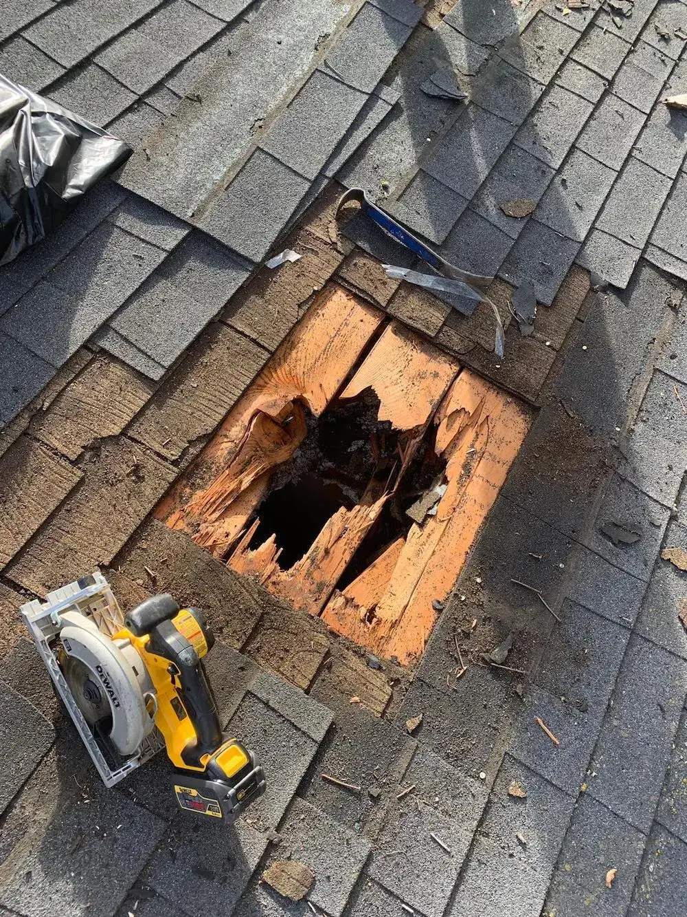 damaged roof section with asphalt shingles removed, exposing rotted plywood and a large hole, surrounded by roofing tools and debris during repair work